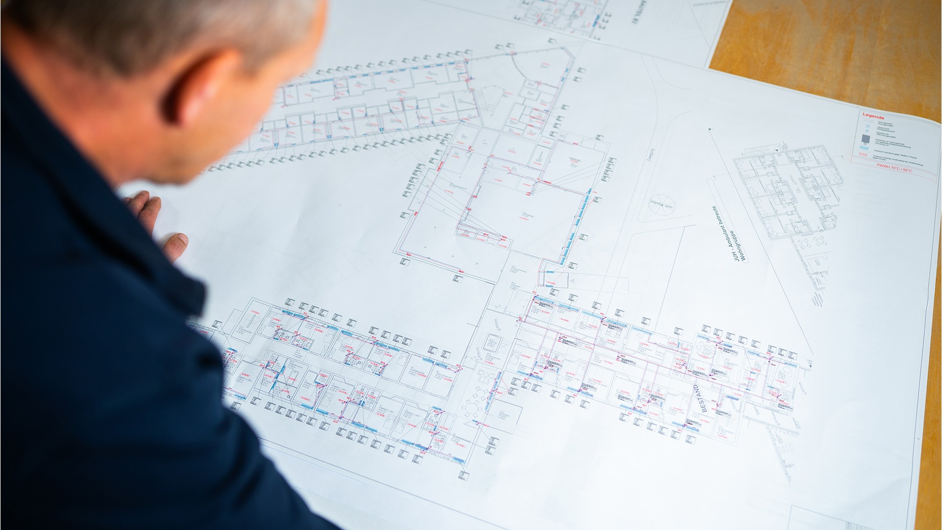 Eine Person beugt sich über einen großformatigen, detaillierten Grundrissplan eines Gebäudekomplexes.